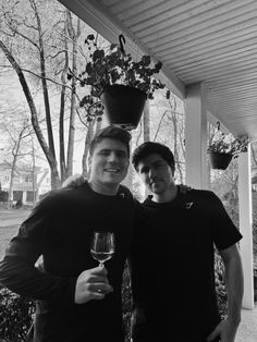 two men standing next to each other holding wine glasses in front of a porch with potted plants