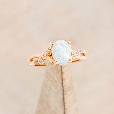 a white opal and diamond ring sitting on top of a piece of wood in front of a beige background