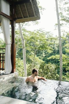 a man sitting in the middle of a large hot tub with trees behind him and white curtains hanging over it