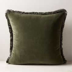 a green velvet pillow with fringe trim on the front and back, sitting against a white wall