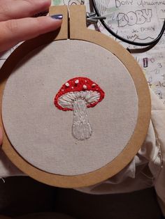 a person holding up a small embroidery project with a red mushroom on it's side