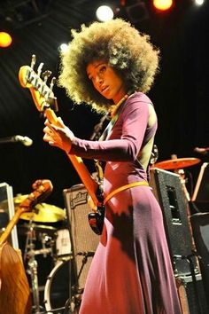 a woman in a purple dress plays an electric guitar on stage with other instruments behind her