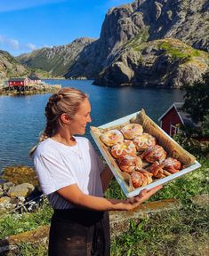 a woman holding a box full of pastries in front of a lake and mountains