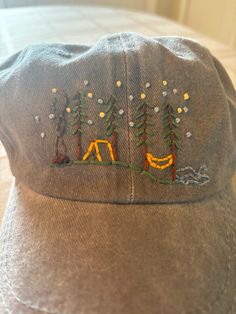 a gray hat with embroidered camping scene on the front and trees in the back, sitting on top of a bed