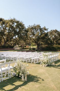 an outdoor ceremony setup with white chairs and flowers in vases on the grass, surrounded by trees