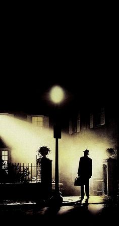 a man walking down a street at night in the fog with a hat and coat on