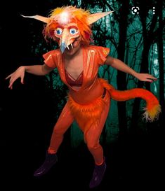 a person dressed in an orange costume with horns and big blue eyes is standing in front of some trees