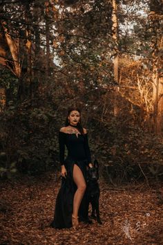 a woman in a black dress standing next to a dog on a leaf covered ground