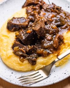 a white plate topped with meat and grits next to a fork on top of a wooden table