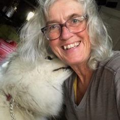 an older woman is hugging her white dog's face and smiling at the camera