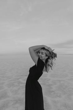 a woman in a black dress standing on the beach with her arms behind her head