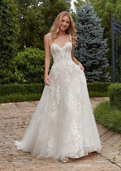 a woman in a white wedding dress standing on a brick walkway with trees and bushes behind her