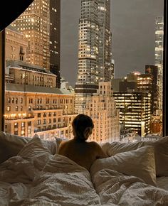 a woman is sitting in bed looking out the window at the cityscape and skyscrapers