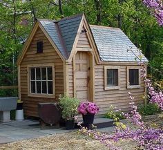 a small wooden house in the middle of some trees and bushes with purple flowers around it