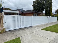 a white fence in front of a house with grass and trees on the side walk