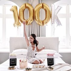 a woman sitting on top of a bed in front of gold balloons and desserts