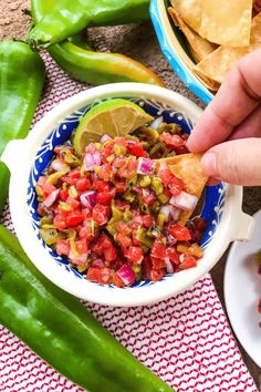 a person dipping salsa into a bowl with tortilla chips and green peppers on the side