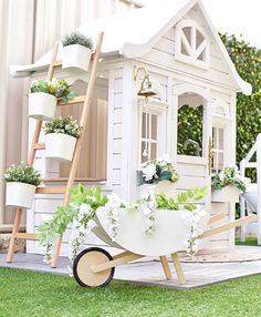 a small white house with flowers in the wheelbarrow and potted plants on top