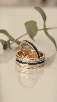 A custom wedding ring set from Staghead Designs sits on a white tile background. This 14K yellow gold set features our "Rio" design wedding band with a centered lapis lazuli inlay & two outer mother of pearl inlays. The other ring is our narrow "Eterna" band featuring a lapis lazuli inlay. This fully customizable set can also be made in 14K white or rose gold & with a wide variety of unique inlay materials including wood, antler, turquoise, or even materials you send in! Lapis Wedding Ring, Whiskey Barrel Wedding Ring, Antler Wedding Rings, Matching Wedding Ring Sets, Matching Wedding Ring, Handcrafted Engagement Ring, Wood Wedding Ring, Mens Gold Wedding Band, Matching Wedding Rings