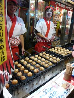 two women in red aprons cooking food on an outdoor grill with buns and muffins