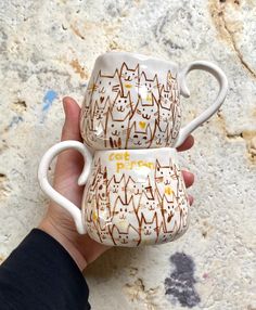 two coffee mugs with designs on them are held in someone's left hand