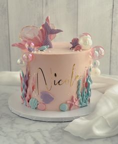 a pink cake with mermaid decorations on top and name written on the side, sitting on a white plate