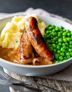 two sausages, mashed potatoes and peas in a white bowl on a table