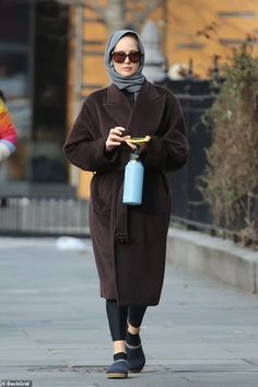 a woman walking down the street with a blue water bottle in her hand and wearing a brown coat