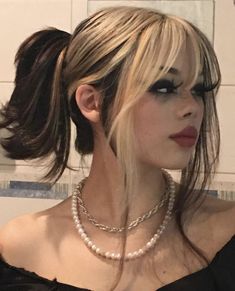 a woman with long hair wearing a black top and pearls on her neck, in front of a tiled wall