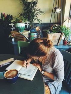 a woman sitting at a table writing on a piece of paper next to a cup of coffee