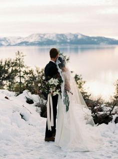 a bride and groom standing in the snow looking out over water with mountains in the background