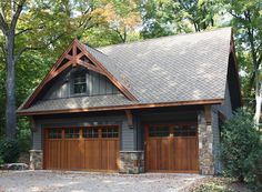 a two car garage with stone and wood accents on the roof, surrounded by trees