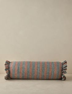 an orange and blue striped pillow with fringes on the bottom, sitting against a beige wall