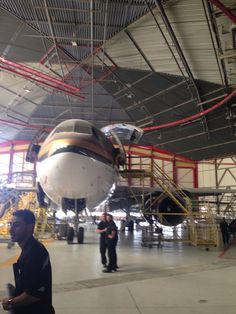 two men are standing in front of an airplane inside the hanger with other people around it