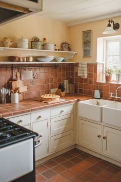 Terracotta tiles set the stage in this classic English cottage kitchen.

The warm tones are enhanced by open shelves showcasing rustic pottery.

Dual farmhouse sinks and wooden countertops create a practical workspace while maintaining the kitchen’s timeless appeal.