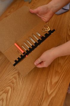 a pair of hands holding a brown box with scissors and pencils in it on top of a wooden table