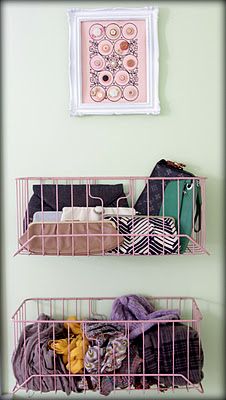 two pink wire baskets filled with clothes next to a wall hanging on a white wall