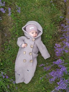 a baby is laying in the grass wearing a gray coat and hat with buttons on it