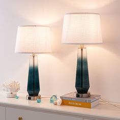 two lamps sitting on top of a white dresser next to a book and lamp shade