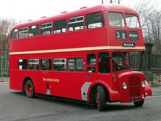 a red double decker bus driving down a street next to a tall metal fence and trees