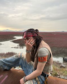 a woman with dreadlocks sitting on the ground next to a river and holding her hand up to her face