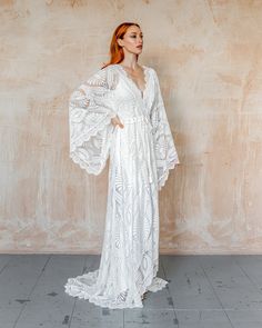 a woman standing in front of a wall wearing a long white dress with ruffled sleeves