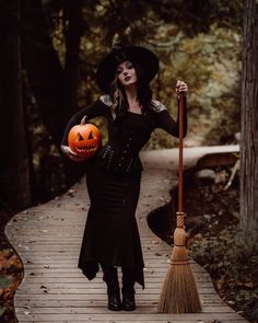 a woman dressed as a witch with a broom and pumpkin on a path in the woods