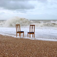 two wooden chairs sitting on top of a sandy beach next to the ocean and crashing waves