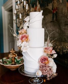 a three tiered white wedding cake with flowers on the top and bottom, sitting on a table in front of a fireplace