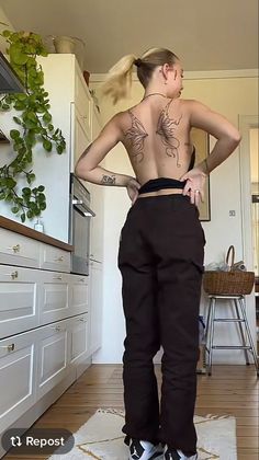 a woman with tattoos on her back standing in a kitchen and looking at the floor