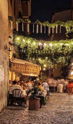 people sitting at tables in an alleyway with plants growing on the walls and hanging lights above them