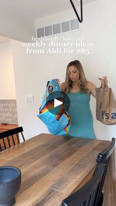 a woman is holding up some bags on the table in front of her, while standing next to a dining room table