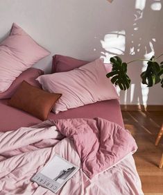 an unmade bed with pink sheets, pillows and a book on the floor next to a potted plant