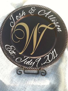 a black and gold wedding plate with the names of two people on it, sitting on a white cloth